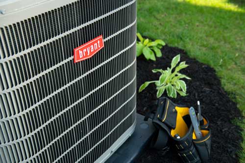 Close view of a Bryant outdoor heat pump by a mulched garden bed; a yellow-and-black tool belt with pliers rests at the base, indicating service work