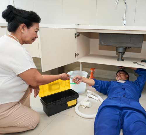 Plumber in blue uniform fixing the pipes of a sink while the customer hands them a tool