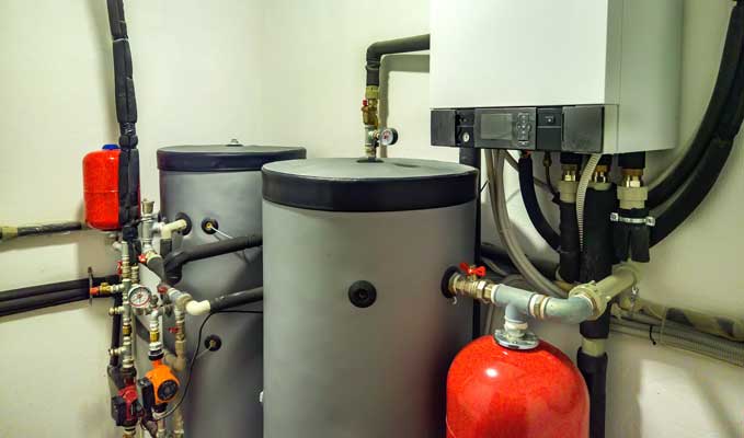 Residential boiler room with a wall-mounted condensing boiler, two insulated hot-water tanks, red expansion tank, and piping.